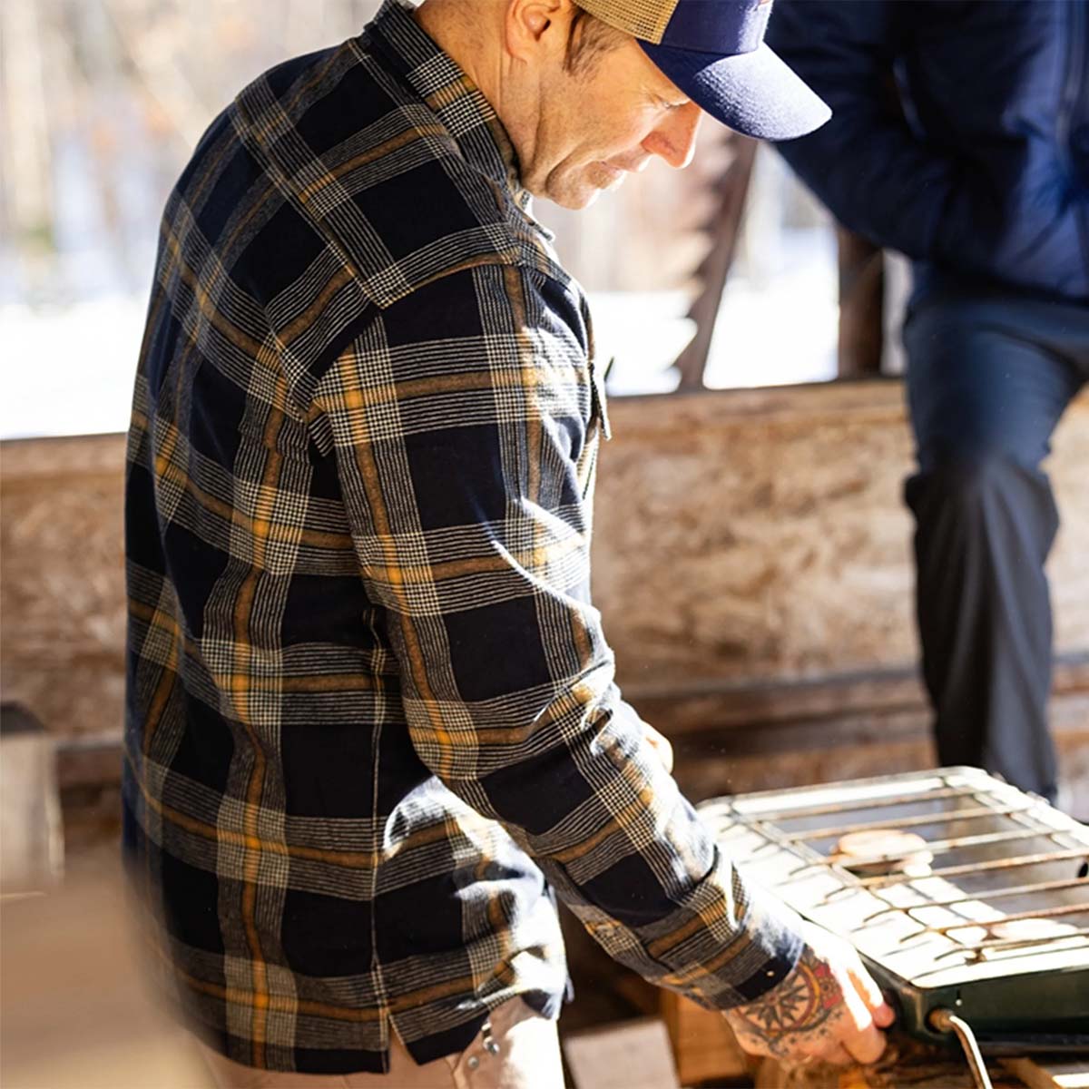 A man wearing a navy blue and yellow plaid flannel shirt and a baseball cap, working at a small outdoor grill in a rustic setting.