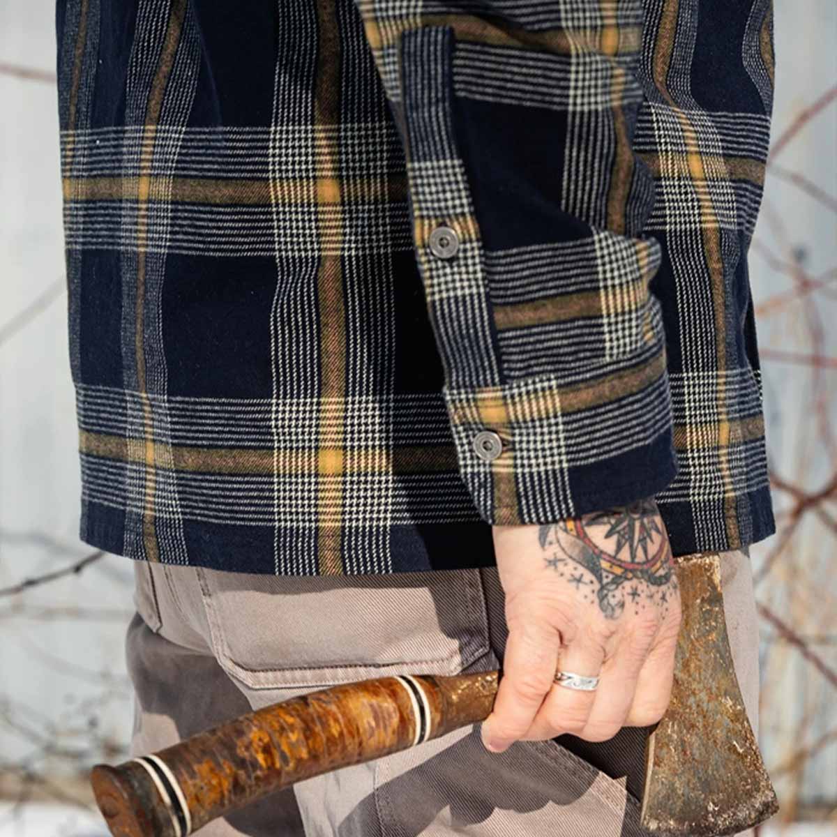 A close-up of a person wearing a navy blue flannel shirt with a yellow and white plaid pattern, holding a rustic wooden-handled axe outdoors.
