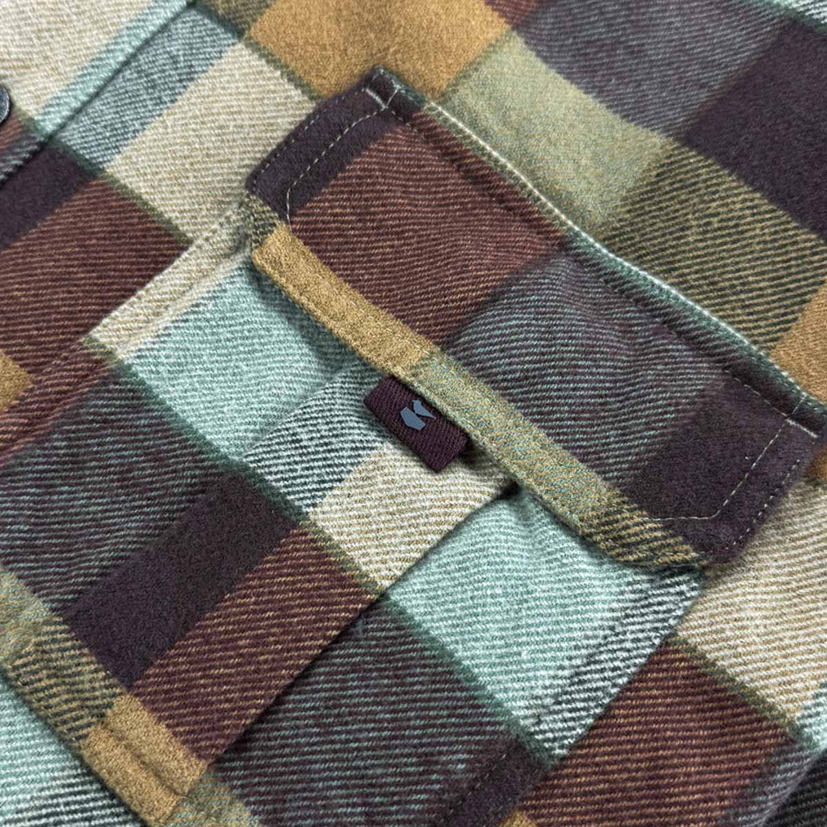 Close-up of a plaid fabric garment, likely a flannel shirt or jacket, showing a pocket flap with a small blue embroidered logo and a color-blocked pattern in muted green, brown, beige, and dark plum tones.