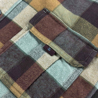 Close-up of a plaid fabric garment, likely a flannel shirt or jacket, showing a pocket flap with a small blue embroidered logo and a color-blocked pattern in muted green, brown, beige, and dark plum tones.