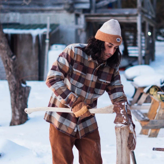 A person in a snowy outdoor setting wearing a brown and beige plaid flannel shirt, brown pants, tan gloves, and a cream and orange beanie hat while holding a wooden log with an axe.
